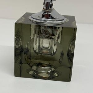 May include: A vintage lighter with a chrome top and a square, olive-green glass base. The base has a series of circular indentations on each side. The lighter is a classic design, suitable for a collector or as a decorative item.