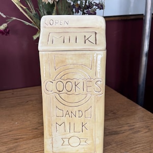 May include: A beige ceramic cookie jar in the shape of a milk carton. The jar features the words "COOKIES AND MILK" and "OPEN MILK" in brown. It is placed on a wooden surface.