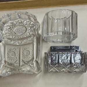 Pode incluir: Um cinzeiro de vidro lapidado transparente, um pequeno recipiente cilíndrico de vidro e um isqueiro de vidro lapidado são exibidos. O cinzeiro tem um design de estrela. O isqueiro tem uma parte superior de metal prateado.