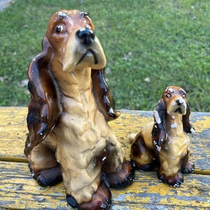 May include: Two ceramic dog figurines, one larger than the other, both brown and white with long ears. The larger dog is sitting with its head tilted up, while the smaller dog is sitting with its head tilted down.