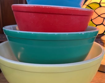 May include: A stack of four vintage mixing bowls in various colors: light blue, red, teal, and yellow. The bowls are nested inside each other, showcasing their different sizes and hues. The bowls have a classic, rounded shape.