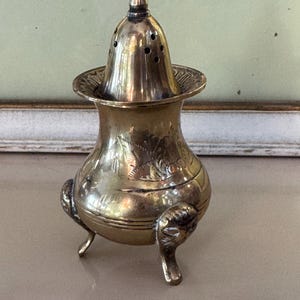 May include: A vintage brass incense burner with a perforated lid and three decorative feet. The burner has an ornate design with floral engravings and a small, rounded top. The brass has a warm, aged patina.