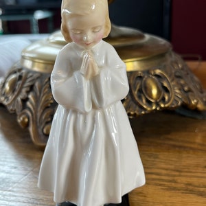 May include: A porcelain figurine of a young girl in a white dress with her hands clasped in prayer. The figurine is standing on a black base.