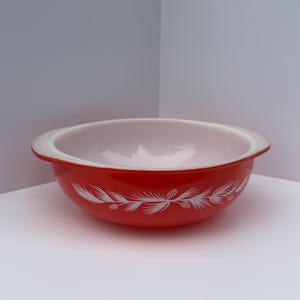 May include: A round, red glass bowl with a white interior and integrated side handles. The exterior features a white graphic pattern of pine needles and pinecones encircling the bowl. The bowl has a smooth, glossy finish and a wide rim.