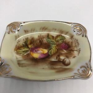 May include: A small, rectangular porcelain dish with a gold rim and a painted design of fruit and leaves. The dish is cream-colored with a brown background.