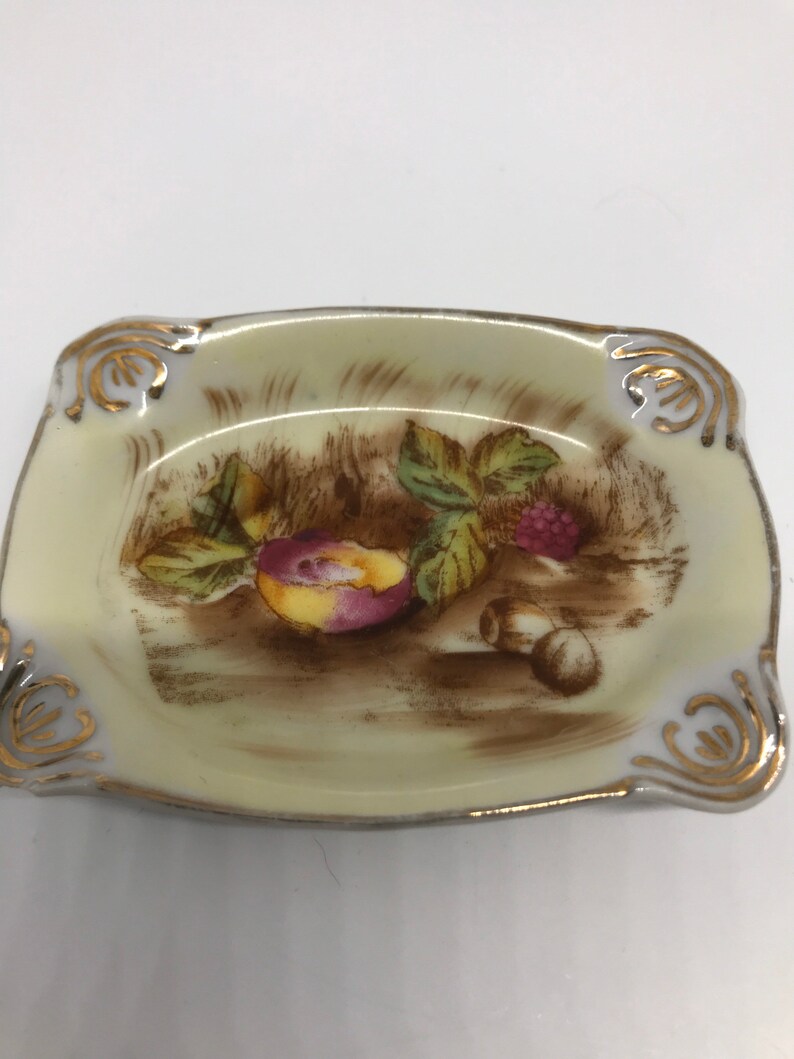 May include: A small, rectangular porcelain dish with a gold rim and a painted design of fruit and leaves. The dish is cream-colored with a brown background.