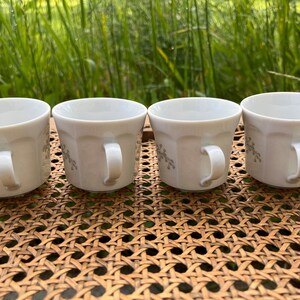 May include: Four white ceramic coffee mugs with a floral design. The mugs are lined up on a woven wicker surface.