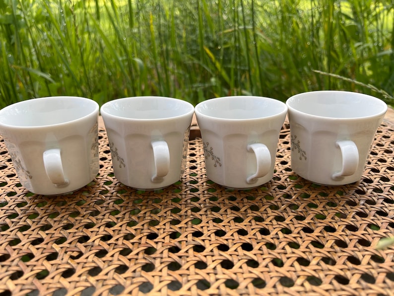 May include: Four white ceramic coffee mugs with a floral design. The mugs are lined up on a woven wicker surface.