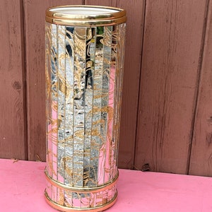 May include: A gold and silver mirrored umbrella stand with a decorative design. The stand has a round base and a cylindrical body.