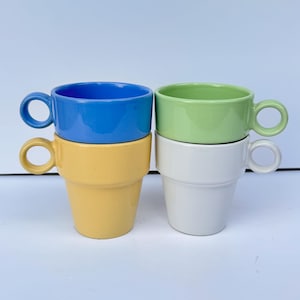 Può includere: Quattro tazze impilate in ceramica blu, verde, gialla e bianca. Ogni tazza ha un manico arrotondato e un corpo cilindrico. Le tazze sono disposte in due pile da due, con i colori alternati.