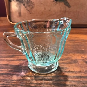 Peut inclure: Un petit pot à crème en verre transparent avec une teinte bleu clair et un motif floral délicat. Le pot à crème a une poignée et une base arrondie.