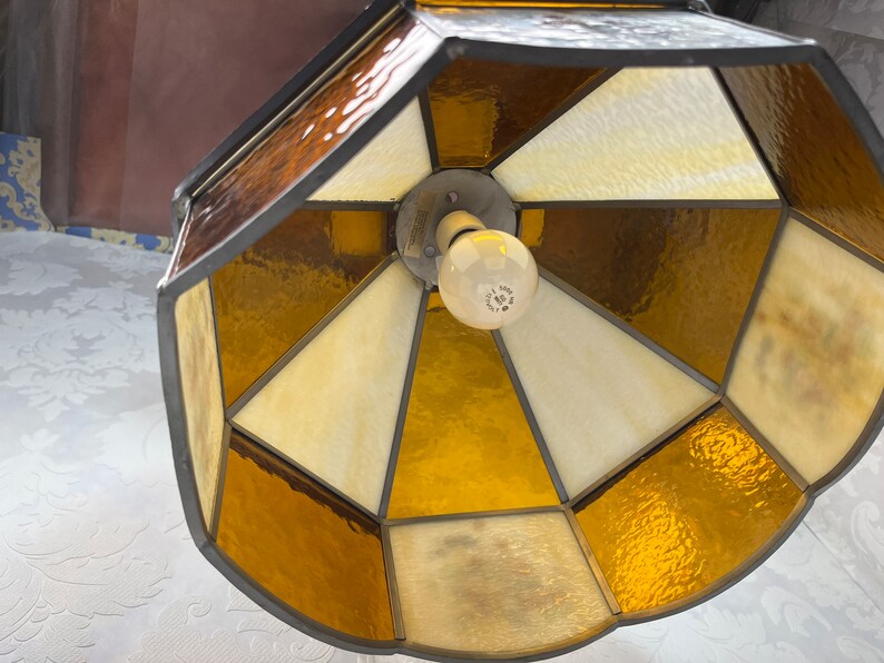 Puede incluir: Una l&aacute;mpara de techo de cristal emplomado con un dise&ntilde;o geom&eacute;trico. La l&aacute;mpara tiene un marco de metal marr&oacute;n y paneles de vidrio transparentes y &aacute;mbar.