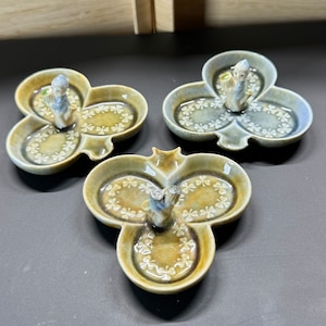 Puede incluir: Tres pequeños platos de cerámica en forma de trébol con un diseño de trébol en relieve. Cada plato tiene una pequeña figura de un duende sosteniendo una olla de oro en el centro.