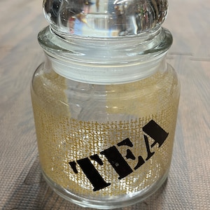 Puede incluir: Un tarro de vidrio transparente con tapa, decorado con una textura similar a la arpillera y la palabra "TEA" en letras negras.