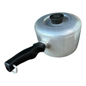 Puede incluir: Una pequeña cacerola de aluminio con tapa y asa negra. La tapa tiene un pomo negro. El asa tiene un agujero para colgar. La cacerola está diseñada para cocinar.