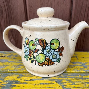 Könnte beinhalten: Eine beigefarbene Teekanne aus Keramik mit einem Blumendesign mit grünen Äpfeln, blauen Blumen und braunen Blättern. Die Teekanne hat eine gesprenkelte Glasur und einen runden Deckel.