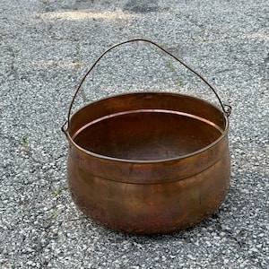 May include: A round copper pot with a polished metallic finish and a curved body. The pot features a thin, arched metal handle attached to two side loops, allowing for easy carrying. The surface shows natural color variations in the copper.