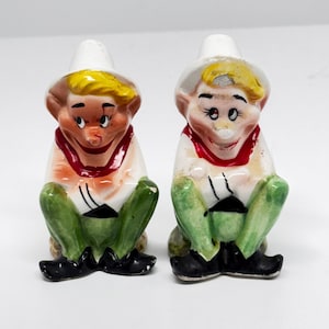 Peut inclure: Une paire de salières et poivrières vintage en céramique. Chaque salière et poivrière représente une figurine assise avec un chapeau blanc, des cheveux jaunes et une écharpe rouge. Les figurines portent des chemises blanches, des pantalons verts et des chaussures noires.