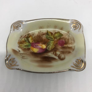 May include: A small, rectangular, cream-colored porcelain dish with a gold trim. The dish features a hand-painted design of a peach, a raspberry, and leaves in brown, pink, and green hues.
