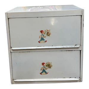 May include: A vintage white metal two-drawer cabinet. Each drawer has a colourful decal of a child carrying flowers. The cabinet has a slightly distressed appearance, suggesting age and use. The design is simple and functional, with decorative decals.