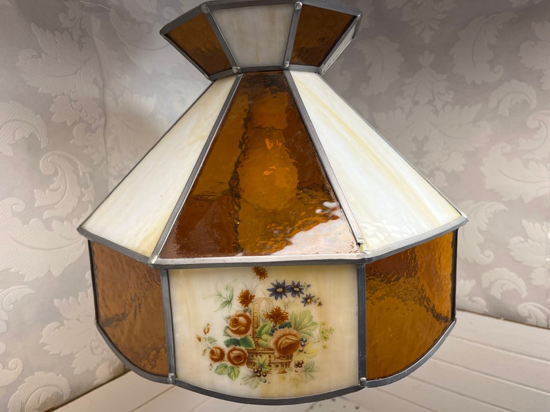 Puede incluir: Una pantalla de l&aacute;mpara de vidrio emplomado con un dise&ntilde;o floral. La pantalla est&aacute; hecha de vidrio transparente y &aacute;mbar y presenta un dise&ntilde;o floral con una cesta de flores. La pantalla tiene forma octogonal y tiene un marco de metal plateado.