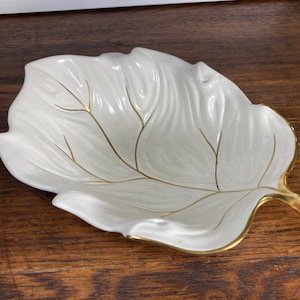 Op de afbeelding: Een wit keramisch bladvormig schaaltje met gouden accenten. Het schaaltje is ontworpen om eruit te zien als een blad met nerven en een steel.