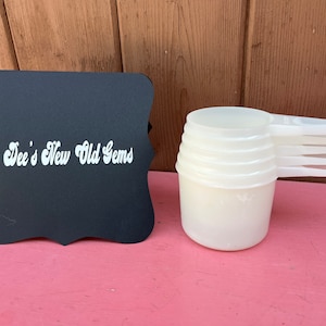 May include: A set of four white plastic measuring cups with handles. The cups are stacked on top of each other. A black chalkboard sign with the text "Dee's New Old Gems" is in the background.