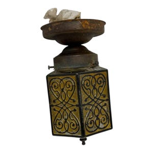 May include: An antique, square-shaped lantern with a decorative, ornate design. The lantern features a dark metal frame with yellow-tinted glass panels. The glass panels are adorned with black, swirling patterns. The lantern has a rusty, aged appearance.