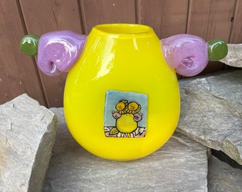 May include: A yellow glass vase with two purple and green handles. The vase has a square tile on the front with a hand-painted design of yellow flowers in a vase.