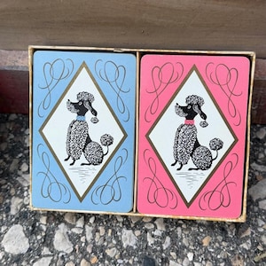 May include: Two decks of playing cards housed in a rectangular box. One deck features a light blue background, and the other features a pink background. Both decks display a white diamond center with a black poodle illustration and gold trim.