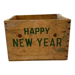 May include: A wooden crate with the words "HAPPY NEW YEAR" printed in green. The crate is rectangular with a natural wood finish and visible wood grain. It has two circular holes and a diamond-shaped logo.