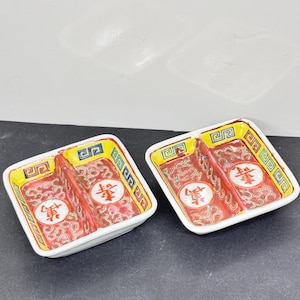 May include: Two square, divided ceramic dishes. Each dish has a red interior with a white central emblem and a yellow border. The rims are white, and the dishes are decorated with a repeating pattern.