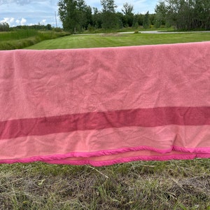 Op de afbeelding: Een gestreept wollen deken in roze en rood met een roze franje die aan een houten hek hangt.