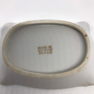 May include: White ceramic ashtray with a rounded oval shape and a slightly raised rim. The bottom of the ashtray has the text "MADE IN OCCUPIED JAPAN" printed in gold.