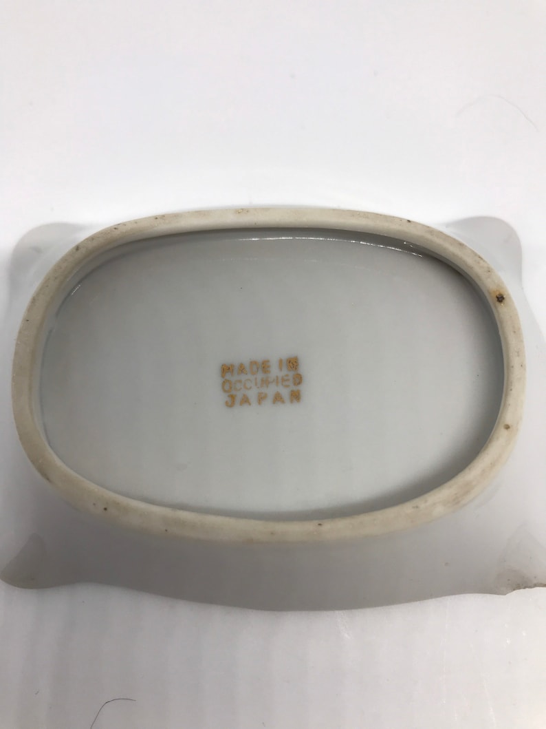 May include: White ceramic ashtray with a rounded oval shape and a slightly raised rim. The bottom of the ashtray has the text "MADE IN OCCUPIED JAPAN" printed in gold.