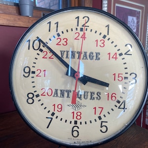 Pode incluir: Um mostrador de relógio branco vintage com números e ponteiros pretos. O mostrador do relógio tem as palavras "Vintage Antiques" impressas. O relógio está em uma moldura circular preta.