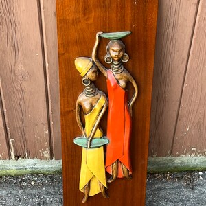 Peut inclure: Une plaque murale en bois avec une sculpture en relief 3D de deux femmes en vêtements colorés. Les femmes portent des robes jaunes et rouges et ont de grandes boucles d'oreilles décoratives. La plaque est de style moderne du milieu du siècle.