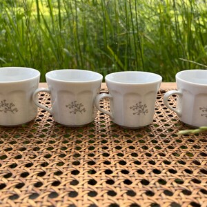 May include: Four white ceramic coffee cups with a floral design. The cups are sitting on a woven wicker surface.