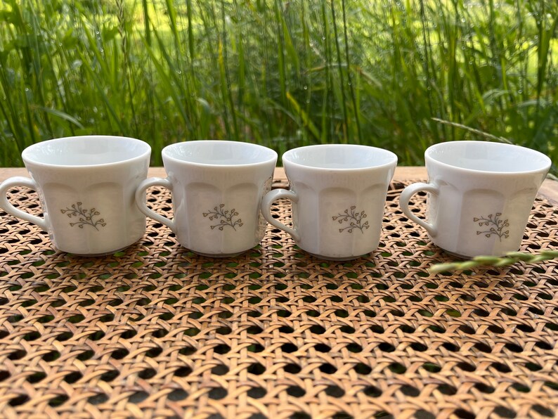 May include: Four white ceramic coffee cups with a floral design. The cups are sitting on a woven wicker surface.