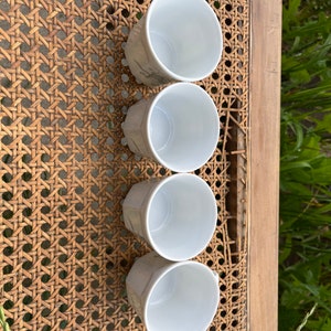 May include: Set of four white ceramic cups with a subtle pattern. The cups are arranged in a row on a woven surface.