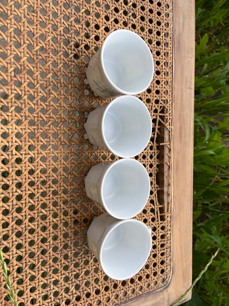 May include: Set of four white ceramic cups with a subtle pattern. The cups are arranged in a row on a woven surface.