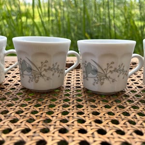 May include: Four white ceramic coffee mugs with a floral design in shades of brown and green. The mugs have a rounded shape and a handle on the side.