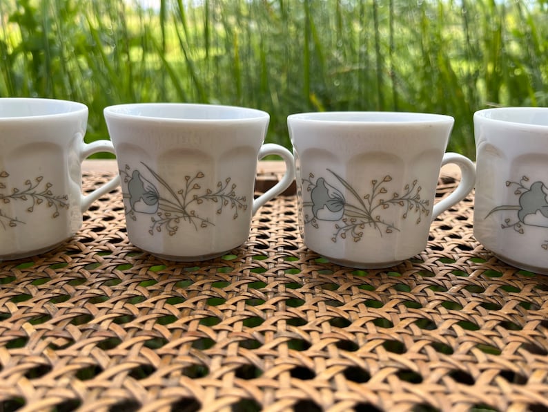 May include: Four white ceramic coffee mugs with a floral design in shades of brown and green. The mugs have a rounded shape and a handle on the side.