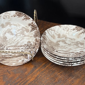 Può includere: Set di sei ciotole per zuppa in ceramica marrone e bianca con un design di scena di campagna. Le ciotole sono impilate l'una sull'altra.