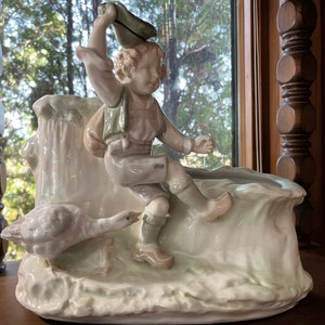 May include: A white porcelain planter shaped like a waterfall with a boy figurine holding a hat and a goose figurine. The planter has a green and brown glaze.