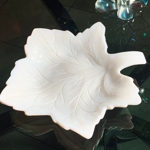 Puede incluir: Un plato de vidrio blanco opaco con forma de hoja con un diseño de hoja detallado en relieve. El plato está sobre una superficie negra.