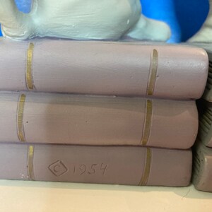 May include: A stack of three lavender-colored ceramic books with gold-colored vertical lines on the spines. The bottom book has the year "1954" and a copyright symbol etched into the side. The books are part of a larger decorative piece.