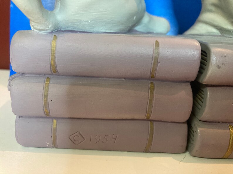 May include: A stack of three lavender-colored ceramic books with gold-colored vertical lines on the spines. The bottom book has the year "1954" and a copyright symbol etched into the side. The books are part of a larger decorative piece.