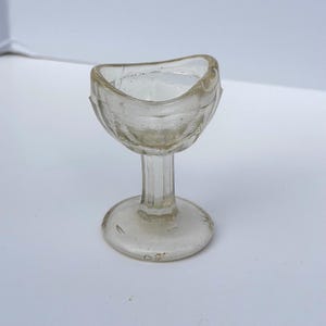 Puede incluir: Una copa lavaojos de vidrio transparente con un borde contorneado diseñado para ajustarse a la cuenca del ojo, un tallo vertical corto y una base circular. El material de vidrio es transparente con burbujas de aire y un tallo moldeado.