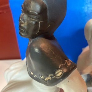 May include: A ceramic figurine featuring a black-skinned bust with a dark gray head and shoulders. The figurine has a white base and is set against a blue and red background. The bust is a side profile.